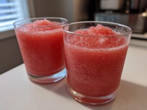 Close-up of two glasses filled with bright red, icy strawberry daiquiri mixture sitting on a white countertop.