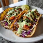 Two perfectly grilled shrimp taco shells filled with seasoned shrimp, purple cabbage slaw, and drizzled with a creamy green sauce.