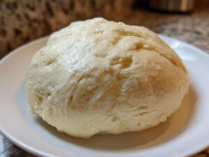 A close-up shot of a freshly made, smooth ball of fufu resting on a white plate.