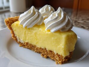 A close-up of a thick slice of bright yellow lemon pie with a graham cracker crust, topped with swirls of whipped cream.