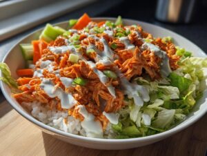 A vibrant buffalo chicken bowl featuring shredded orange buffalo chicken over white rice, topped with ranch dressing, lettuce, carrots, and celery.