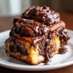 Close-up of gooey chocolate cinnamon rolls stacked on a white plate, dripping with thick chocolate glaze.