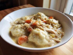 A close-up of a white bowl filled with creamy chicken and dumplings, featuring shredded chicken and carrots.
