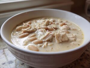 Close-up of a white bowl filled with creamy white chicken chili, showing shredded chicken and white beans.