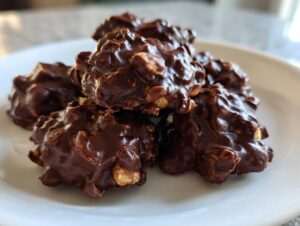 A close-up of several glossy, rich chocolate crockpot peanut clusters piled on a white plate.