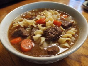 Close-up of a steaming bowl of rich beef noodle soup featuring chunks of beef, egg noodles, and sliced carrots.