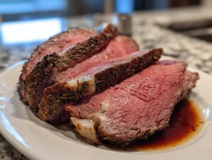 Close-up of three perfectly medium-rare slices of prime rib, crusted with herbs, resting in au jus on a white plate.