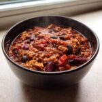 Close-up of a steaming dark bowl filled with rich, thick high protein chili featuring ground meat and kidney beans.