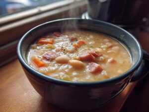 Close-up of a steaming bowl of hearty navy bean soup containing white beans, diced carrots, and ham pieces.