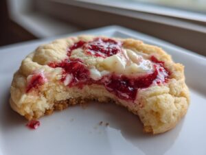 Close-up of a soft strawberry cheesecake cookie with a bite taken out, showing the jam and cream cheese filling.