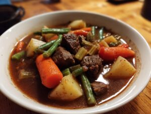 Close-up of a white bowl filled with rich vegetable beef soup, showing chunks of beef, carrots, potatoes, and green beans.