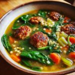 A close-up bowl of flavorful wedding soup featuring browned meatballs, bright green spinach, carrots, and acini di pepe pasta.