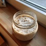 A highly active sourdough starter, showing significant bubbling and foam, sitting in a glass jar on a wooden windowsill.