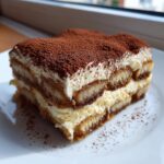 A close-up slice of homemade tiramisu recipe showing layers of cream and coffee-soaked ladyfingers, topped with cocoa powder.