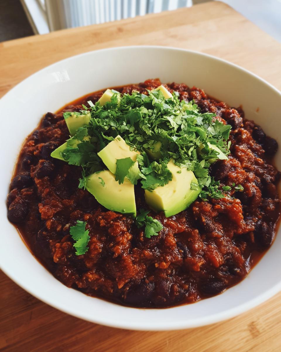 A close-up of a white bowl filled with rich black bean chili, topped with fresh avocado slices and cilantro.