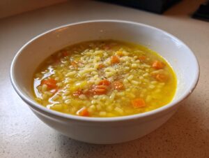 A white bowl filled with hot italian penicillin soup, featuring small pasta, diced carrots, and topped with grated cheese and pepper.