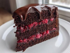 A decadent slice of three-layer chocolate raspberry cake featuring rich dark layers, bright raspberry filling, and a glossy chocolate ganache drip.