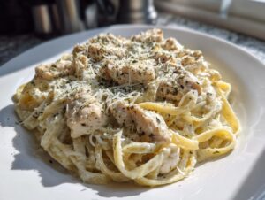 A close-up of a white plate filled with creamy chicken alfredo pasta, topped with diced chicken and grated parmesan cheese.