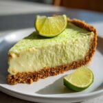 A close-up of a creamy slice of key lime cheesecake on a white plate, topped with lime zest and fresh lime wedges.