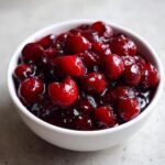 Close-up of glossy, deep red homemade cherry pie filling overflowing slightly in a small white bowl.