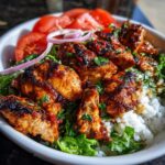 Close-up of a white bowl filled with white rice, green lettuce, sliced tomatoes, and pieces of charred, spicy harissa chicken.