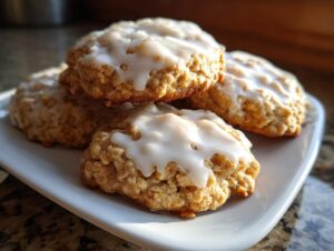 A close-up of several soft, thick iced oatmeal cookies stacked on a white plate, featuring a sweet white glaze.