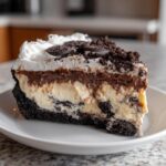 A tall slice of layered no-bake ice cream cake featuring a dark crust, vanilla filling, chocolate layer, and whipped topping with Oreo pieces.