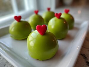Close-up of several shiny, bright green Grinch Oreo Balls topped with small red candy hearts on a white platter.