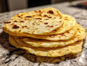 A stack of freshly cooked, soft corn tortillas with light char marks resting on a granite countertop.