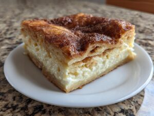 A square slice of sopapilla cheesecake featuring flaky pastry and a thick, creamy filling, topped with cinnamon sugar.