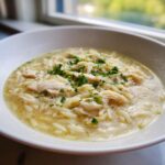 Close-up of a white bowl filled with amazing chicken orzo soup, featuring shredded chicken and topped with fresh parsley.