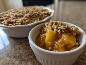 Close-up of a small white ramekin filled with warm, syrupy peaches and topped with crunchy oat crumble from the amazing peach crisp.