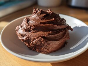 A close-up of a dollop of rich, dark chocolate buttercream frosting swirled high on a white plate.