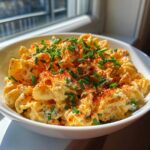 A close-up of creamy deviled egg pasta salad mixed with pasta, topped generously with paprika and fresh chopped chives.