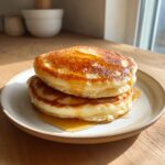 A stack of two fluffy discard pancakes drizzled generously with maple syrup on a light ceramic plate.