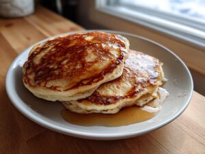A stack of three fluffy discard pancakes drizzled generously with golden syrup on a white plate.