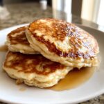 A close-up of three fluffy oatmeal pancakes stacked and drizzled generously with golden maple syrup.