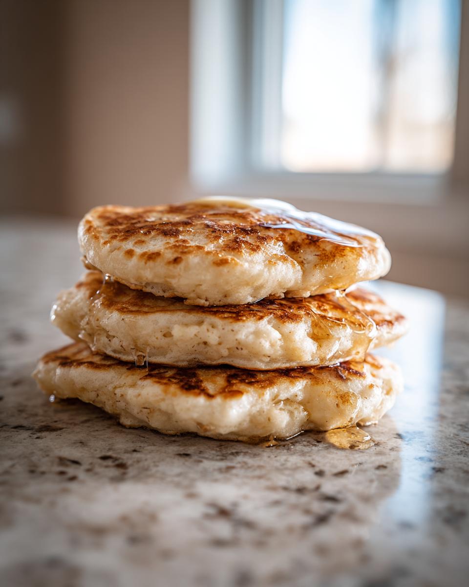 A close-up stack of three fluffy oatmeal pancakes drizzled with syrup, sitting on a granite countertop.