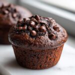 A close-up of a rich, dark chocolate zucchini muffin topped with chocolate chips, sitting on a white surface.