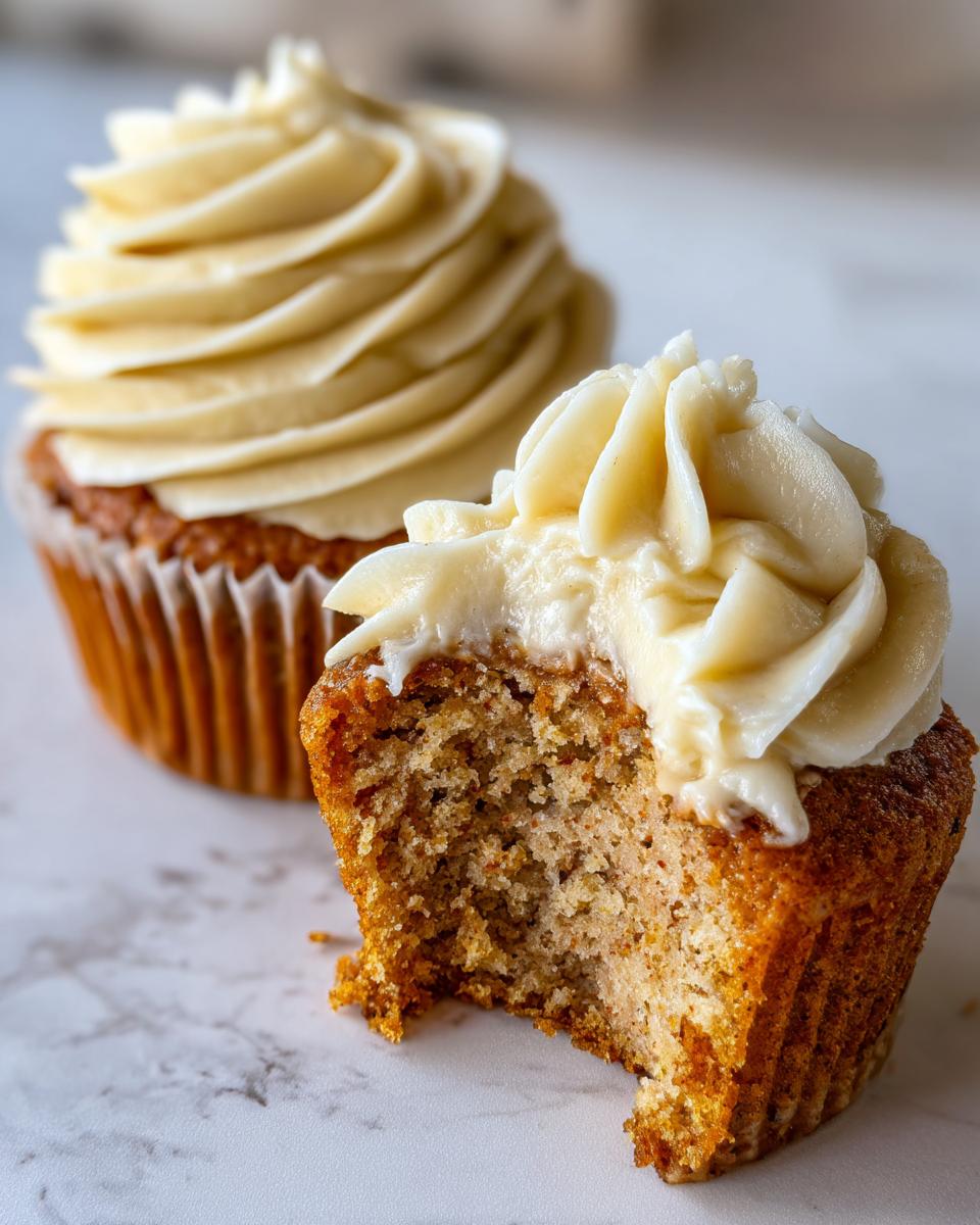 A close-up of a banana cupcake with a bite taken out, showing the moist crumb and thick vanilla frosting.