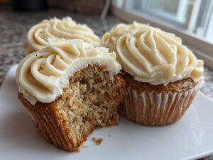 Close-up of moist banana cupcakes with creamy frosting; one has a bite taken out showing the texture.