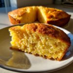 A close-up of a moist, golden slice of cornbread resting on a white plate, with the rest of the cornbread visible in the background.