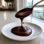 A spoonful of rich, dark chocolate ganache drips onto a white plate, showing its perfect consistency.