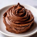A close-up of a perfect swirl of rich, dark chocolate buttercream frosting on a white plate.