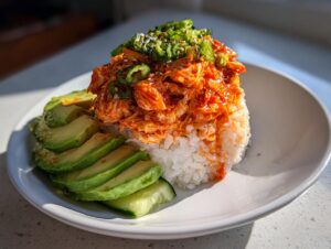 A close-up of a spicy crab sushi bowl featuring bright orange spicy crab mix over white rice, served with sliced avocado and topped with green onions.