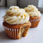Close-up of two banana cupcakes, one with a bite taken out, topped with creamy vanilla frosting.