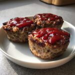 Three perfectly baked mini meatloaf cups topped with a dollop of bright red ketchup glaze, served on a white plate.