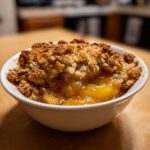 Close-up of a warm serving of peach crisp in a white bowl, showing bubbly fruit filling and a golden brown oat topping.