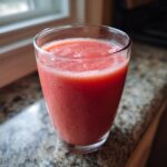 Close-up of a vibrant, pink watermelon smoothie in a clear glass sitting on a granite countertop.