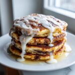A tall stack of golden brown cinnamon roll pancakes generously drizzled with thick white icing.